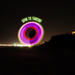 At night, a person spins the glowing Foxtail LED, creating a swirling trail of pink and purple light. Above it, text reads SPIN TO THROW!! City lights shimmer in the background as this exciting light-up night toy glows brightly.