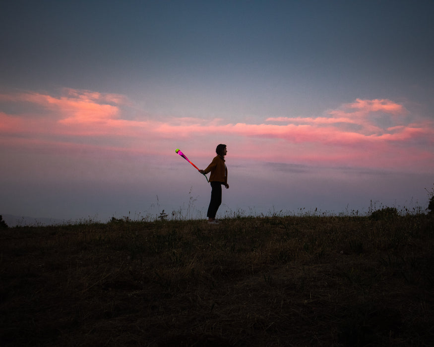 At dusk, a person stands on a grassy hill holding a glowing pink Foxtail LED over their shoulder as the sky darkens and pink clouds streak the horizon.